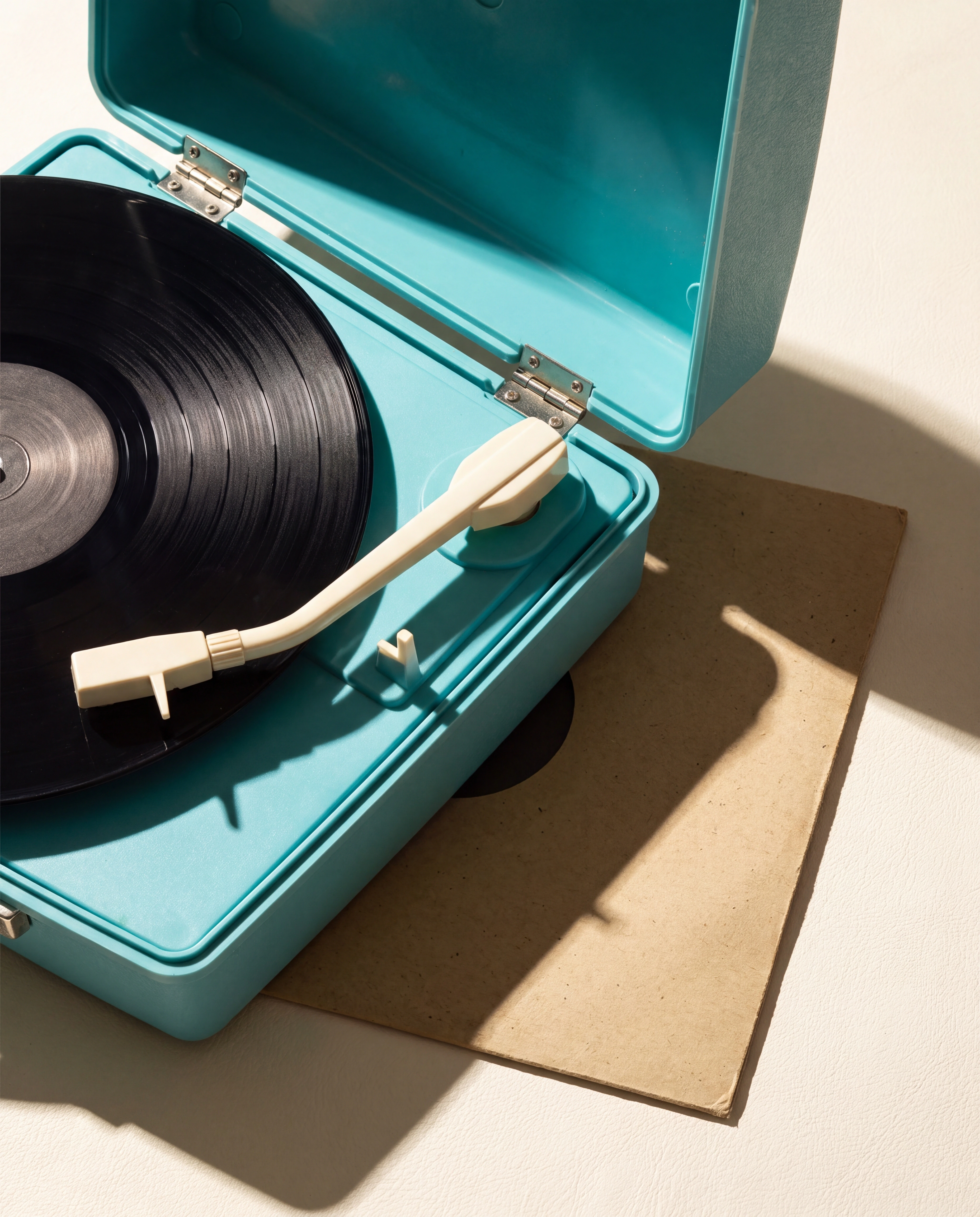 Un tocadiscos portátil turquesa con vinilo negro y packaging color arena bajo el sol.